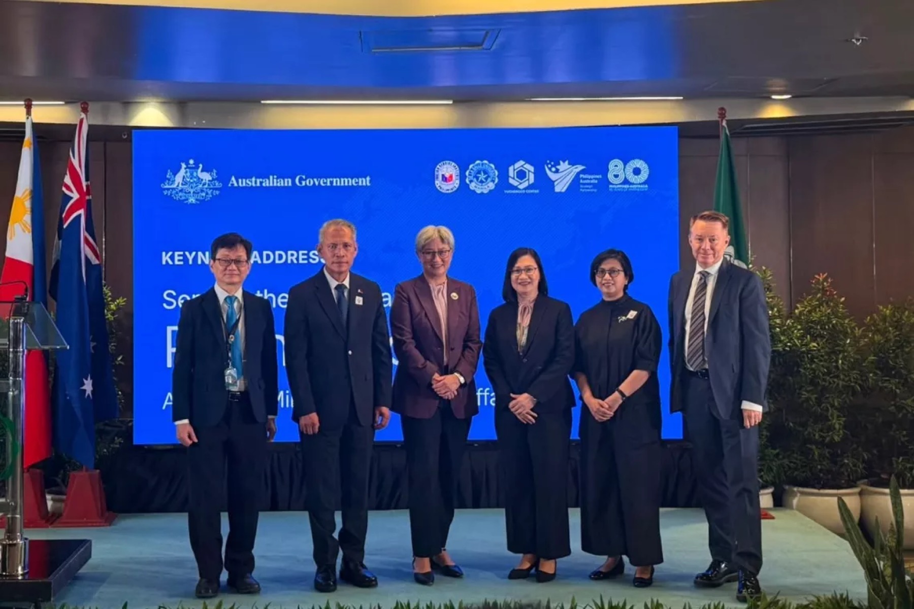 Six people on a low stage between Philippine and Australian flags, before a blue screen carrying the Australian Government crest