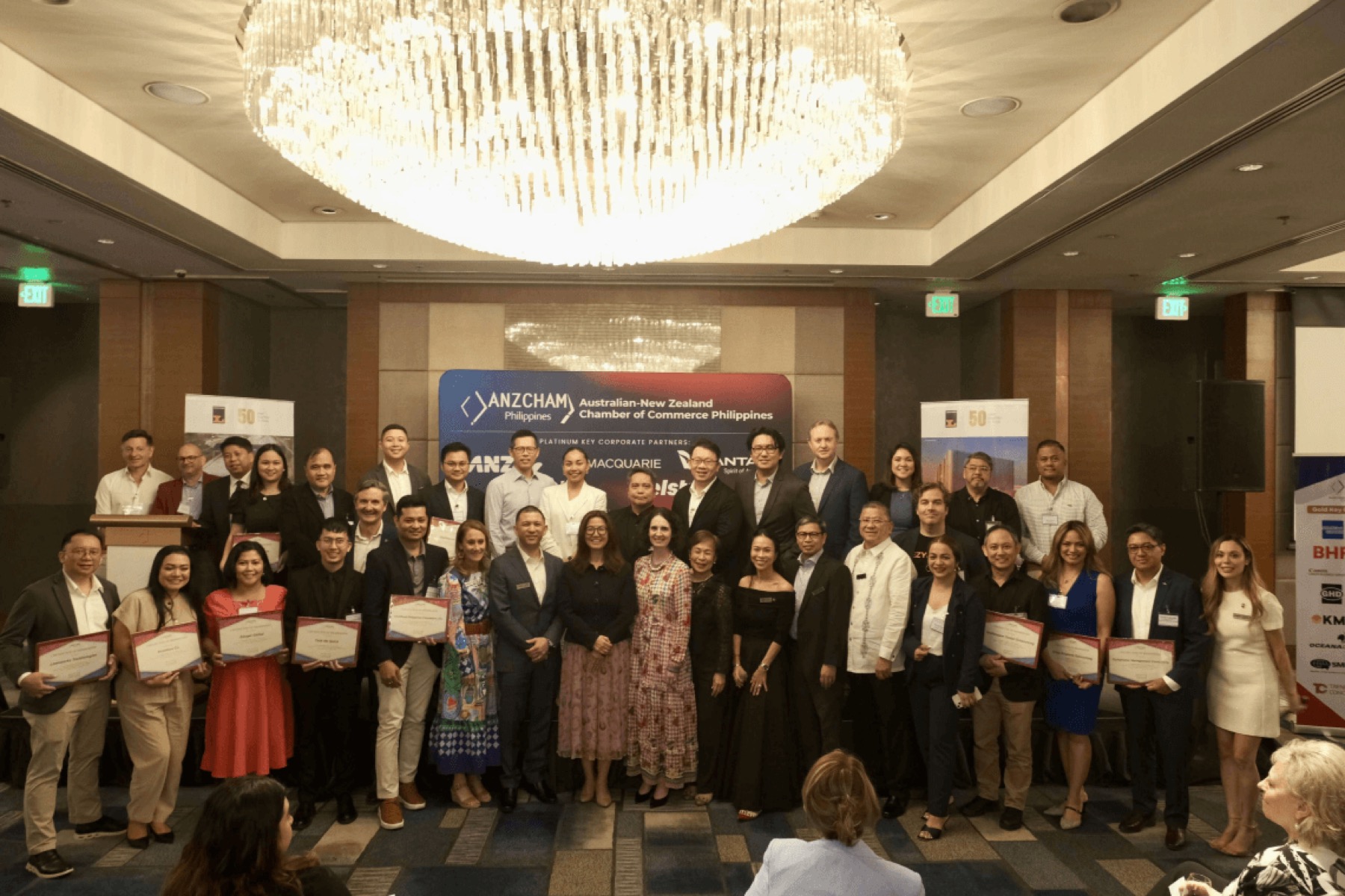 A ballroom under a broad chandelier, with about thirty five guests on stage holding certificates in front of an ANZCHAM Philippines banner