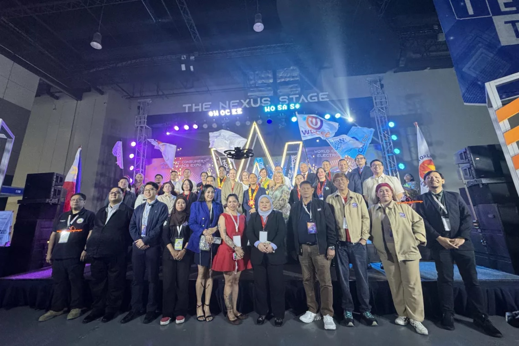 A large group on an exhibition stage under a lit arch and screens reading The Nexus Stage